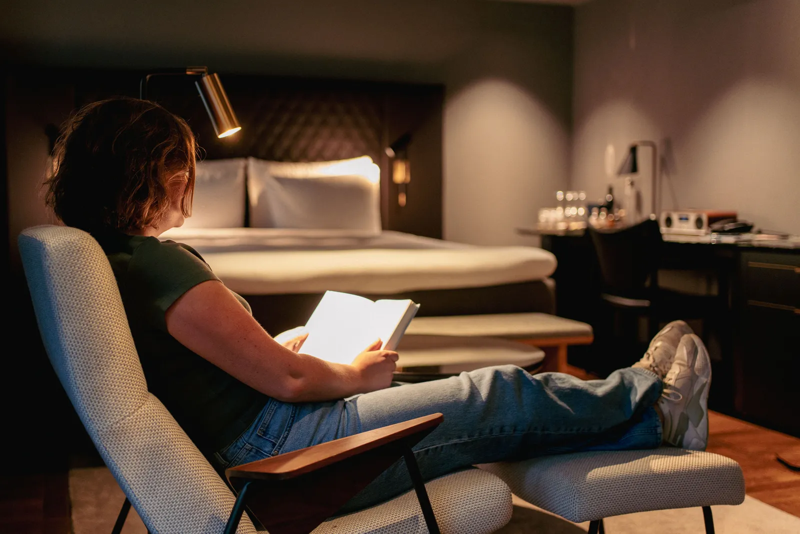 Cliente lisant au fauteuil dans une chambre d’hôtel, espace bureau en arrière-plan